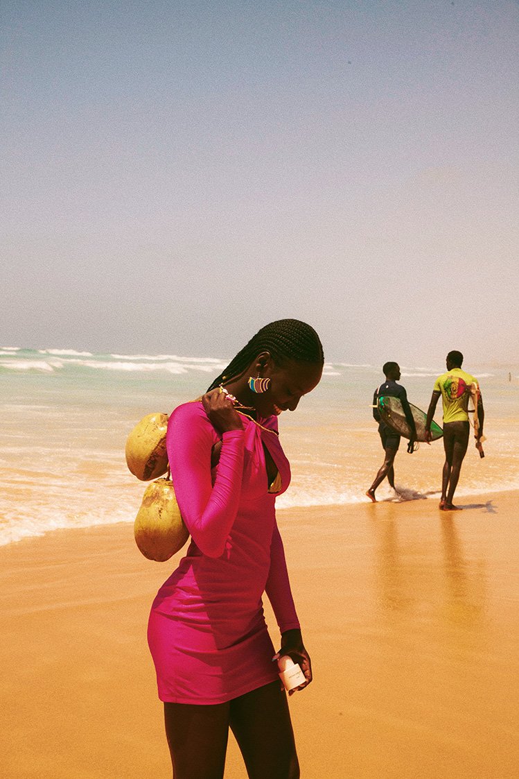 Woman in a pink dress walks on a beach with coconuts, with two surfers in the background, on a sunny day with blue sky and ocean.