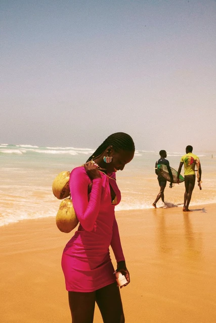 Woman in a pink dress walks on a beach with coconuts, with two surfers in the background, on a sunny day with blue sky and ocean.