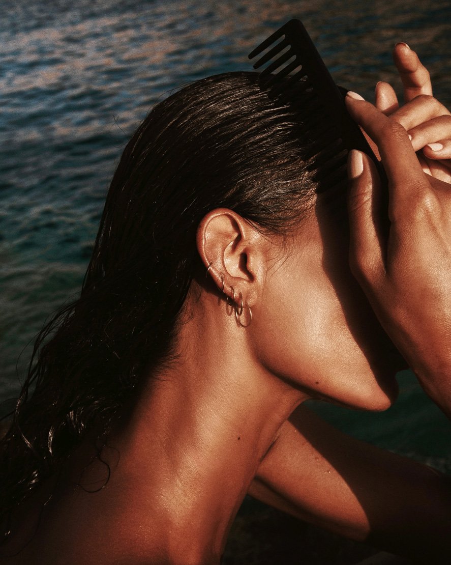 Woman combs wet dark hair with a black comb, against an ocean backdrop. Close-up captures the reflective hair, ear piercings, and hands.