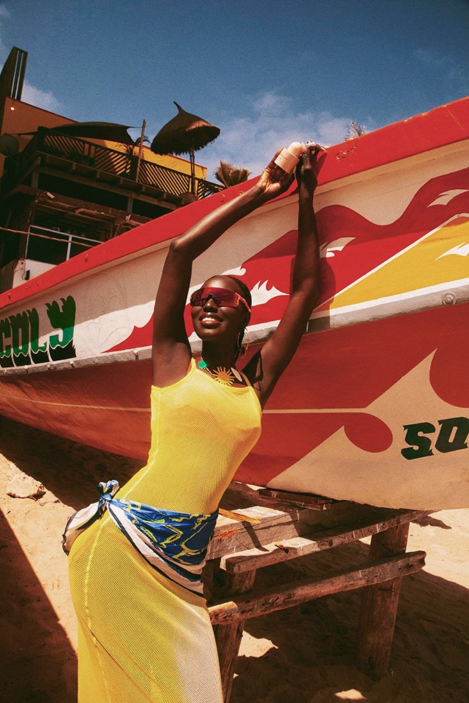 Black woman in a yellow dress posing against a large red and white painted fishing boat on a sandy beach.