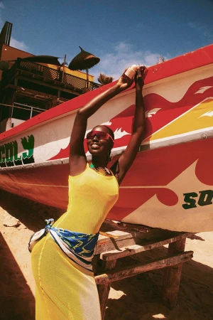 Black woman in a yellow dress posing against a large red and white painted fishing boat on a sandy beach.