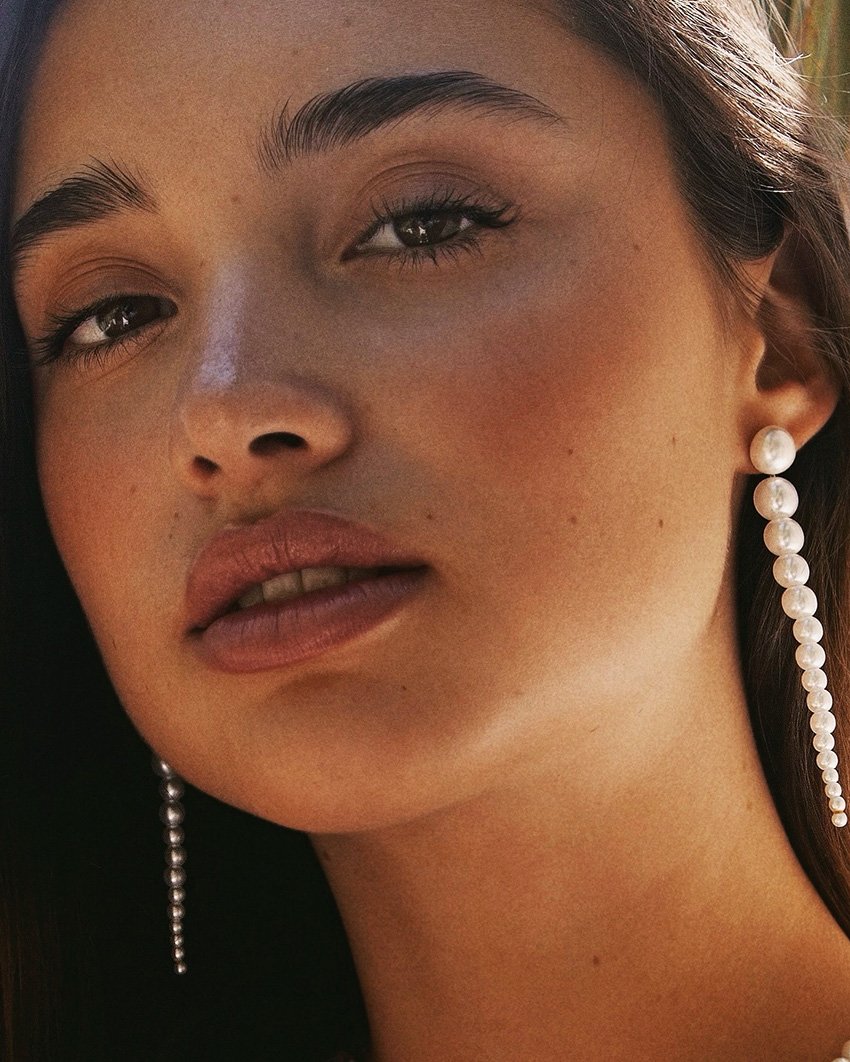 Close-up portrait of a young adult woman with brown hair wearing long pearl earrings, neutral makeup, and soft, diffused lighting.