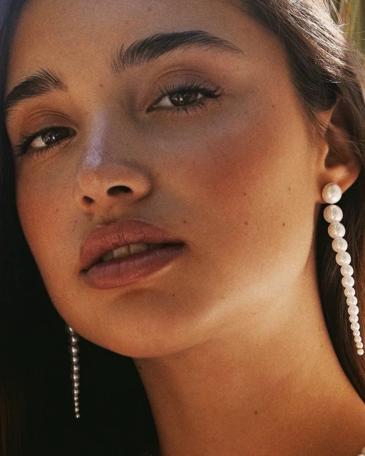 Close-up portrait of a young adult woman with brown hair wearing long pearl earrings, neutral makeup, and soft, diffused lighting.