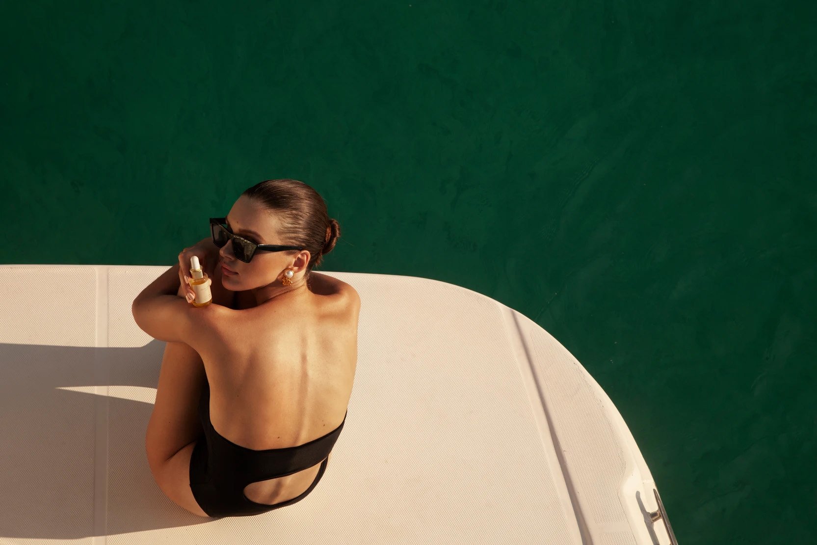 Woman in swimwear applying skin oil on a yacht against a vibrant green water backdrop, with sunglasses and earrings.