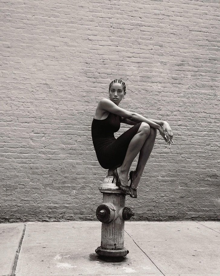 Monochrome portrait of a young woman in a black dress sitting on a fire hydrant in an urban environment with a brick wall behind her.