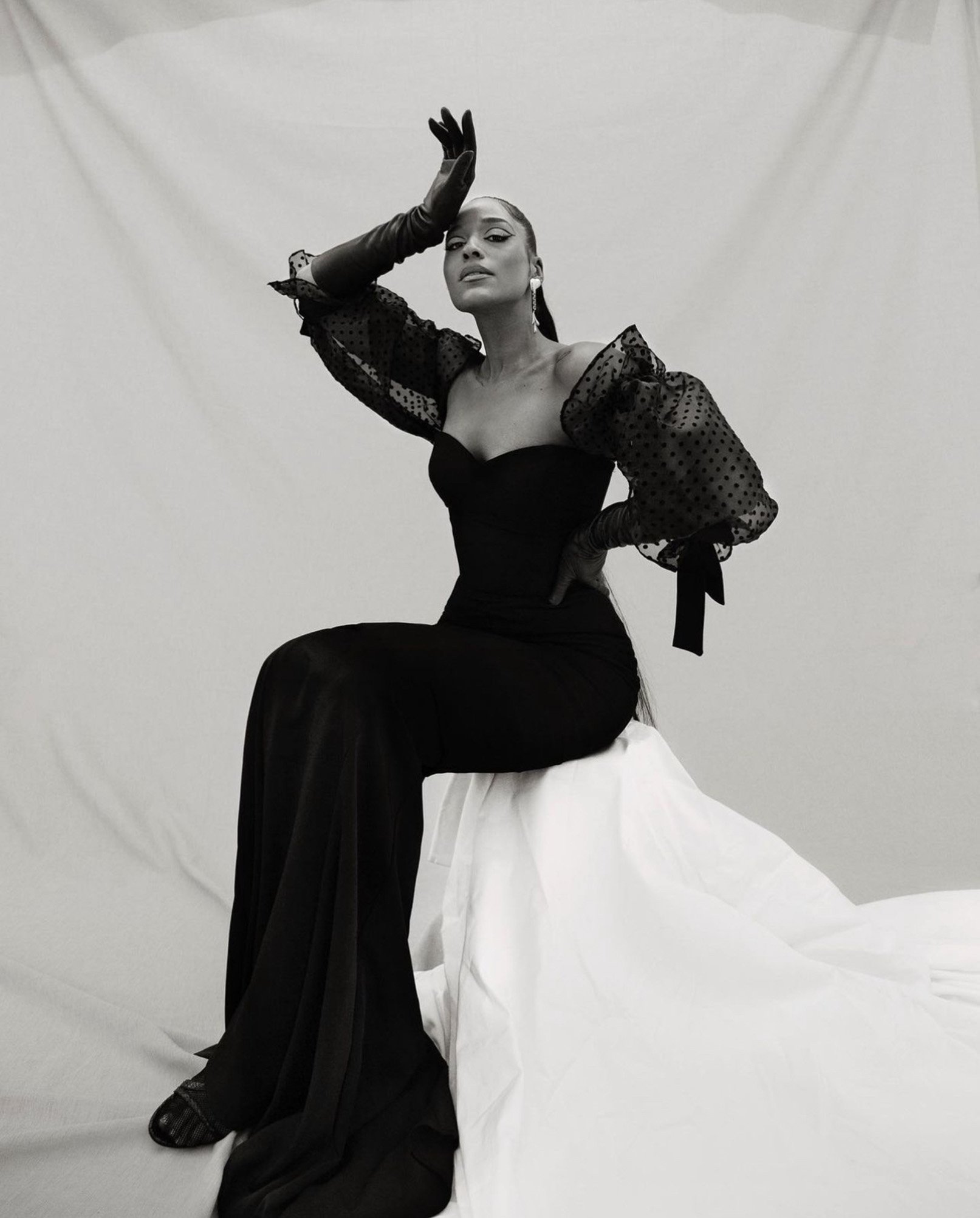 Elegant woman in long black gown with polka-dot sleeves and gloves poses gracefully in a black and white fashion shot.