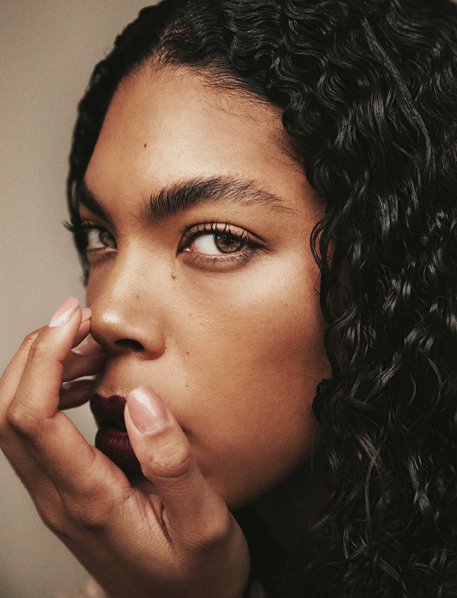 Close-up portrait of a woman with dark, curly hair and deep red lipstick, holding her hand up to her face in an indoor setting.