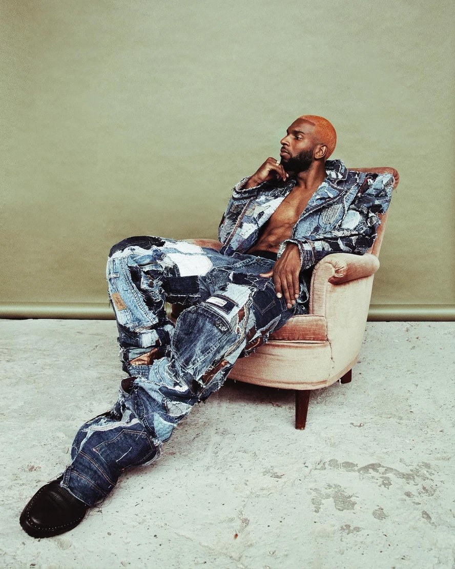 Adult male in patchwork denim suit sits in pink chair against olive-green backdrop with low lighting in a fashion editorial-style shot.