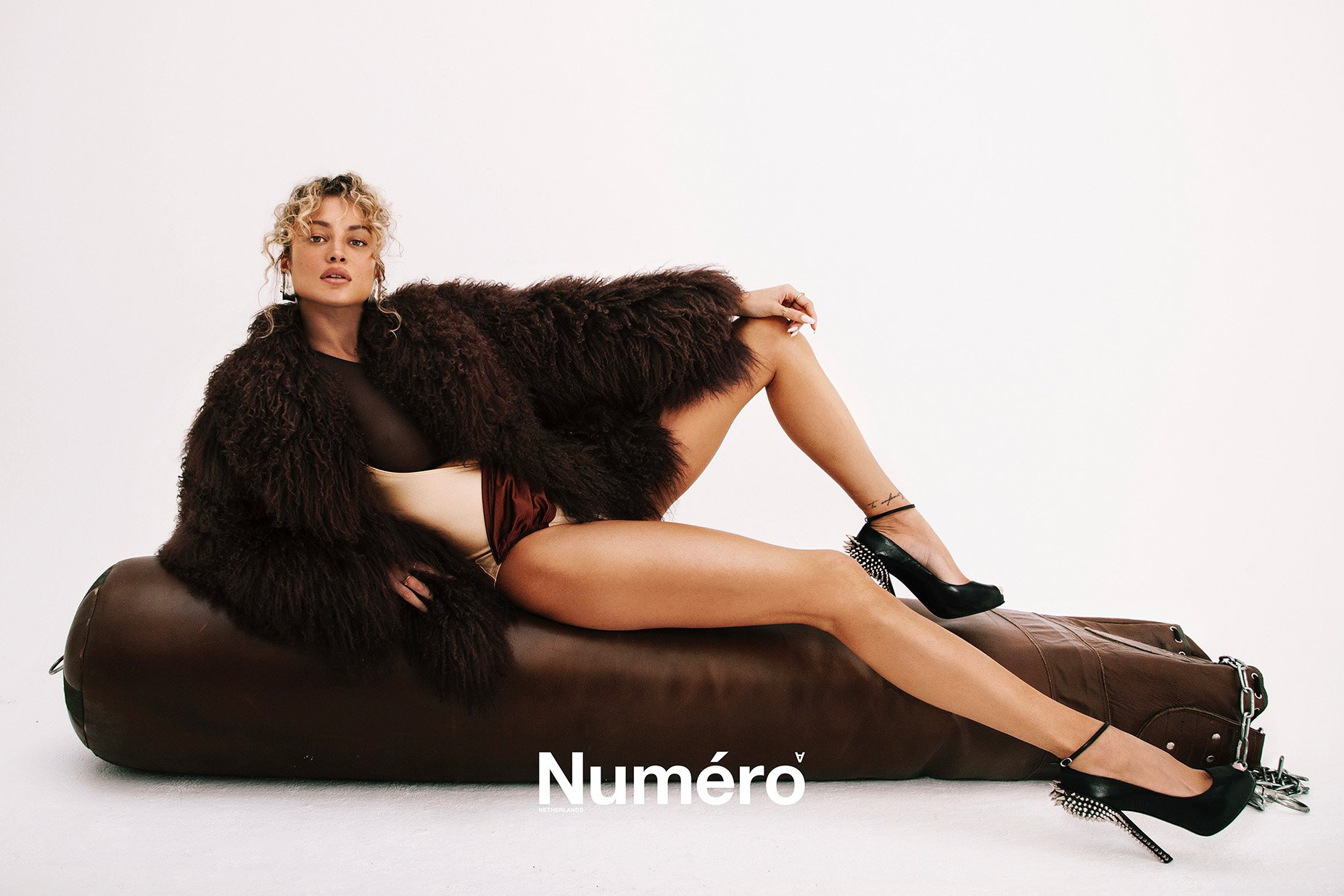 A fashionable woman with blond hair, wearing a fuzzy brown jacket and heels, lounges on a leather ottoman against a white backdrop.