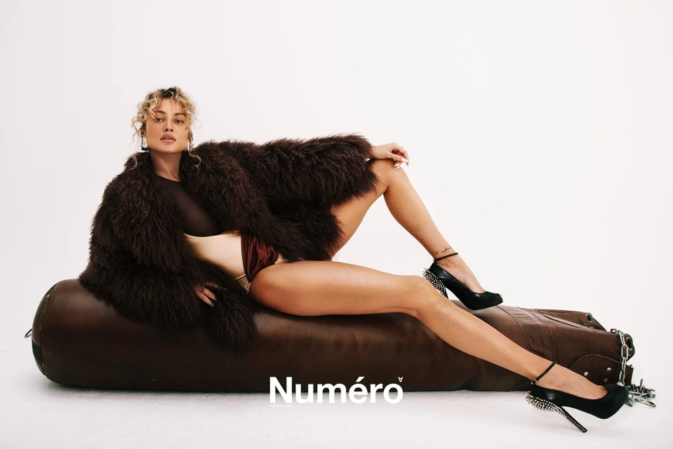 A fashionable woman with blond hair, wearing a fuzzy brown jacket and heels, lounges on a leather ottoman against a white backdrop.