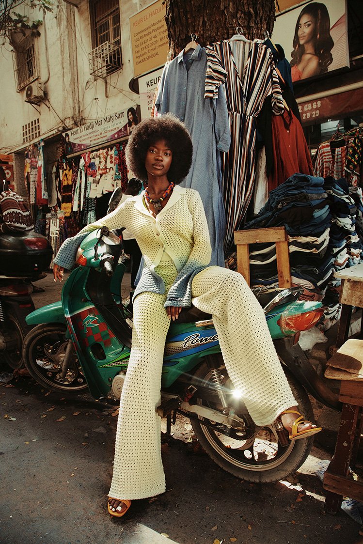 A stylish Black woman with an Afro poses on a green moped in a vibrant outdoor market setting, wearing a cream-colored crochet outfit with tan sandals.