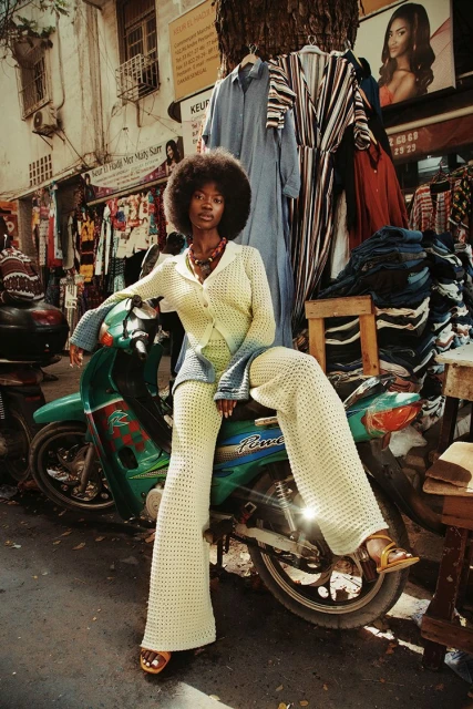 A stylish Black woman with an Afro poses on a green moped in a vibrant outdoor market setting, wearing a cream-colored crochet outfit with tan sandals.