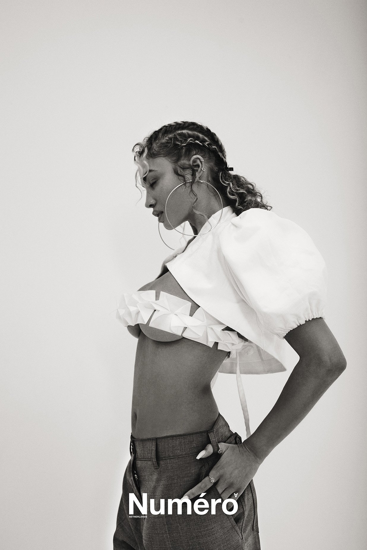 Black and white editorial portrait of a young woman in a cropped top and trousers, wearing jewelry, against a neutral studio background.