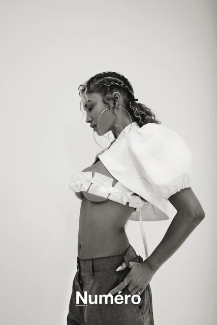 Black and white editorial portrait of a young woman in a cropped top and trousers, wearing jewelry, against a neutral studio background.