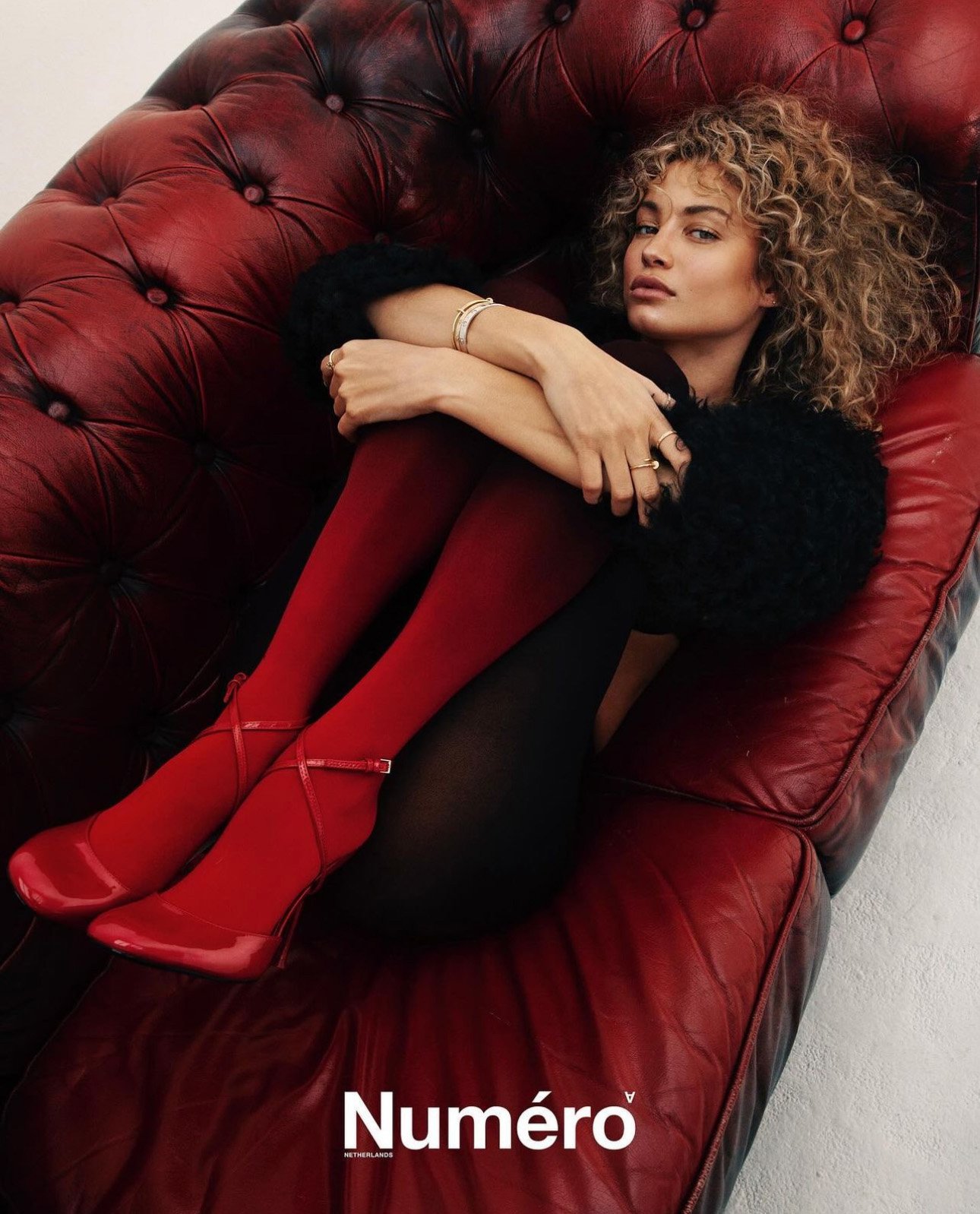 A fashionable adult woman with curly hair reclines on a red leather couch wearing matching red tights and high heels in a studio setting.
