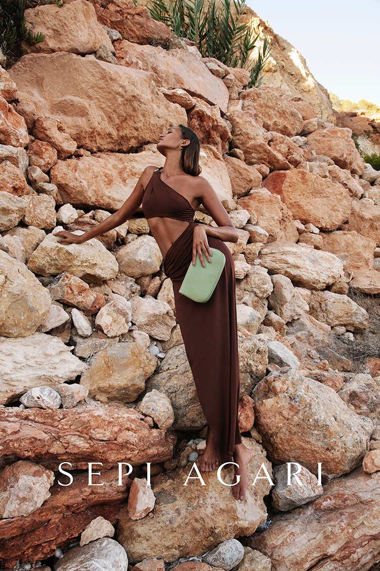 A tan-skinned woman models a brown dress and clutch on a rocky outcrop against a backdrop of tan and brown boulders.