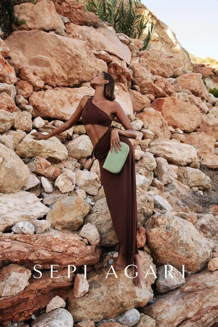 A tan-skinned woman models a brown dress and clutch on a rocky outcrop against a backdrop of tan and brown boulders.