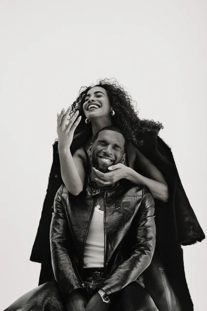 Black and white shot of an African American couple: a woman with curly hair stands behind a seated man in a jacket, both smiling.