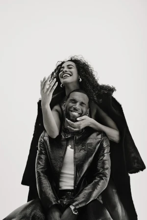 Black and white shot of an African American couple: a woman with curly hair stands behind a seated man in a jacket, both smiling.
