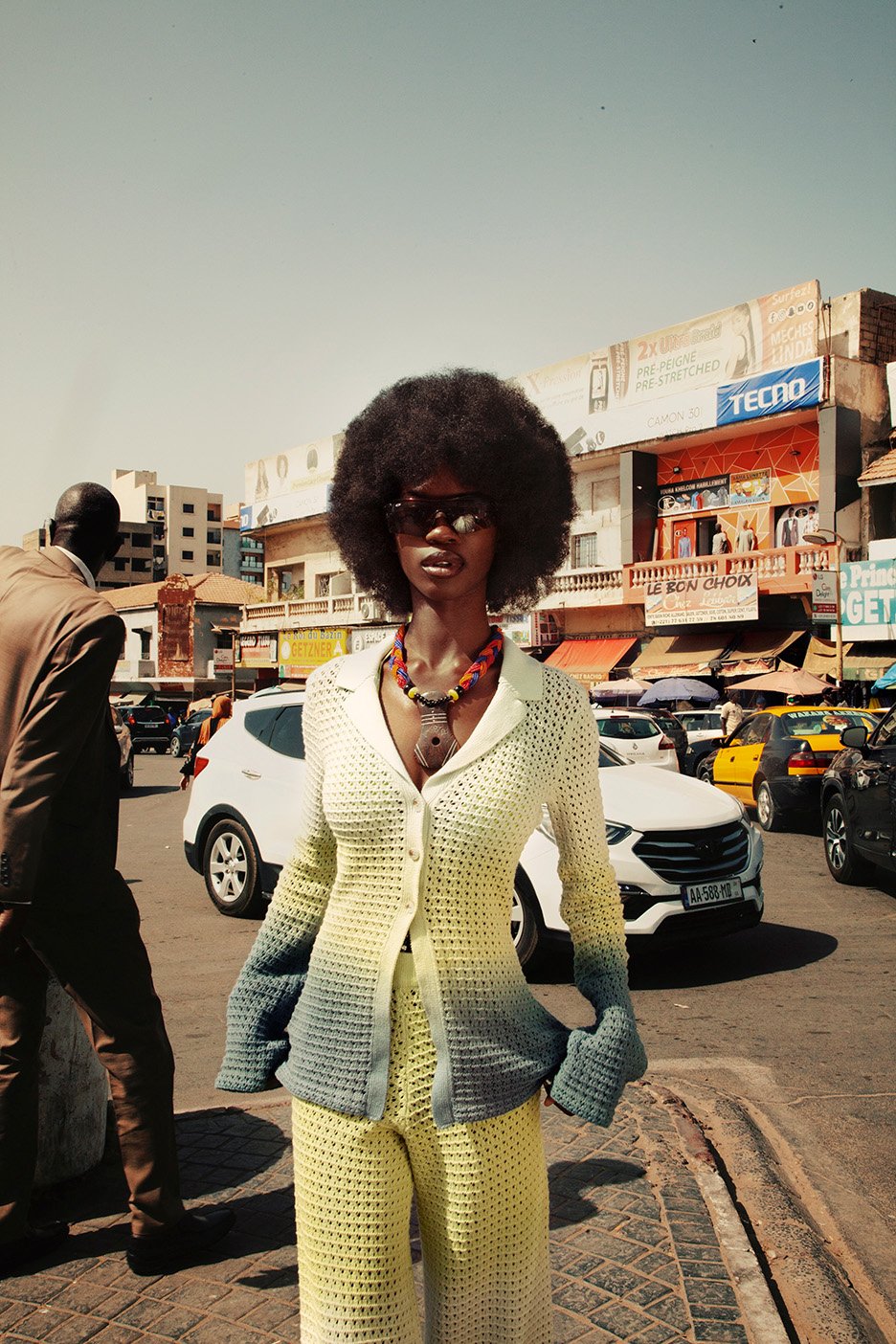 Woman in crocheted suit stands on city street with traffic and buildings in background on a sunny day in an outdoor fashion shot.