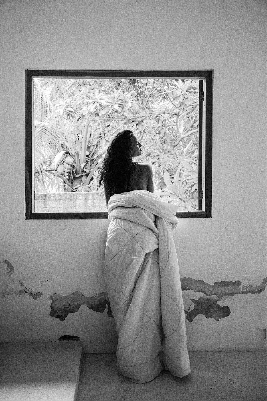 Black and white portrait of a woman gazing through a window draped in a blanket, capturing a serene, intimate moment in an indoor setting.