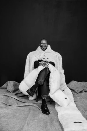 Black man smiling while seated in a chair, draped in an oversized plush white fur coat against a dark backdrop.