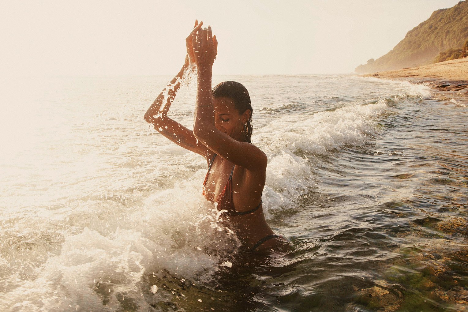 Woman in a bikini standing in the ocean with her hands raised, sunlight reflecting off the water with a landscape in the background.