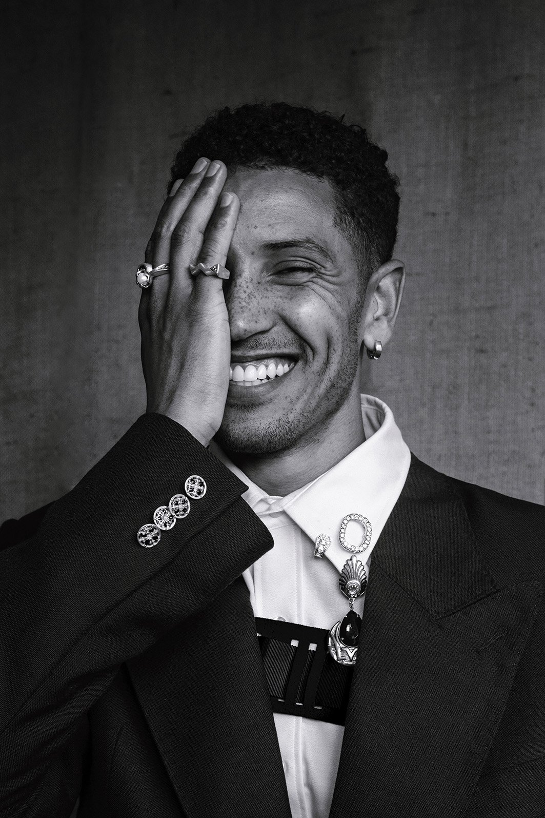 Monochrome portrait of smiling adult male covering one eye with his hand, wearing suit jacket, dress shirt, rings, and earrings.