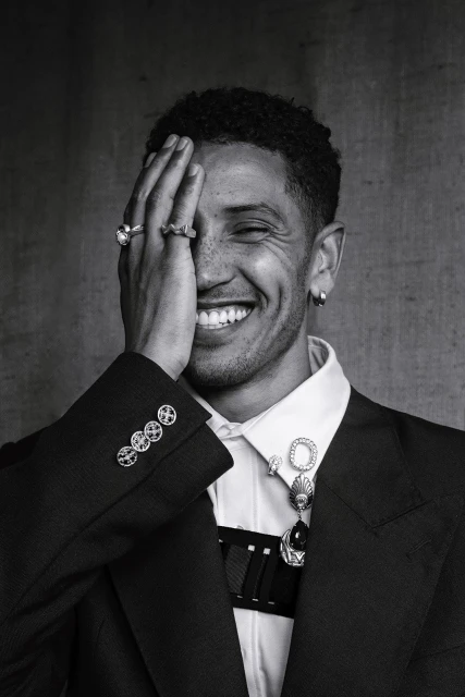 Monochrome portrait of smiling adult male covering one eye with his hand, wearing suit jacket, dress shirt, rings, and earrings.