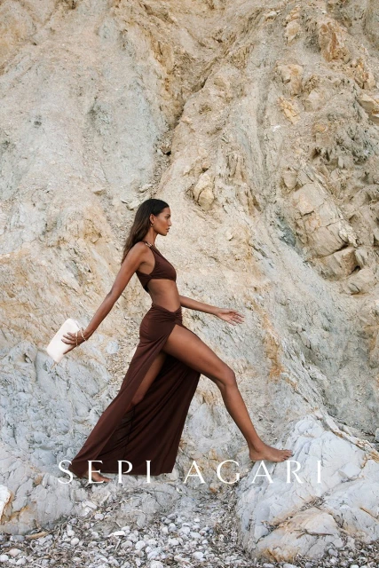 A woman poses on a rocky terrain wearing a brown two-piece outfit with a high slit skirt, accessorized with a bracelet and clutch.
