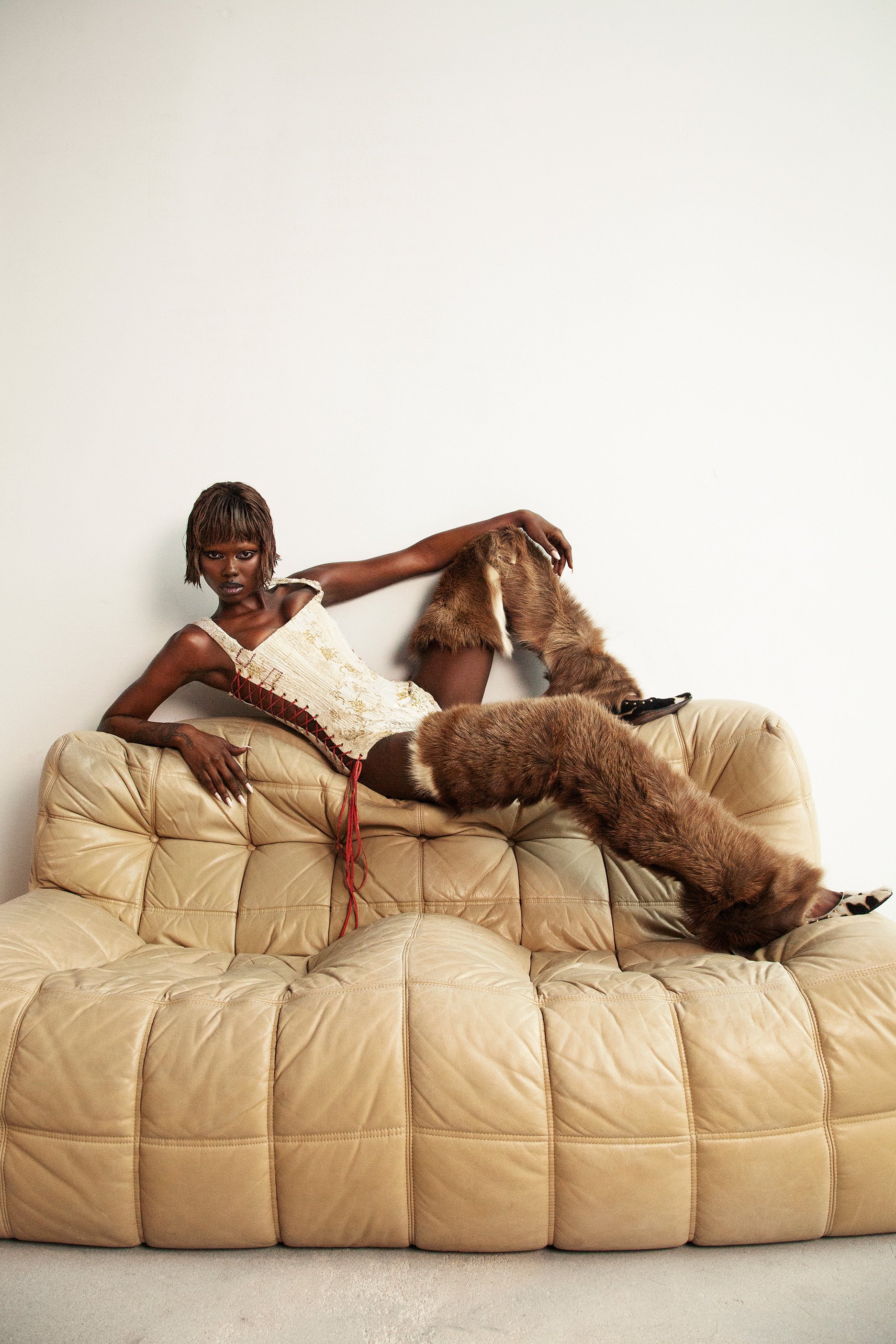 Brunette woman reclines on a tan, tufted sofa wearing a white corset top and furry leg warmers against a white backdrop in a studio.