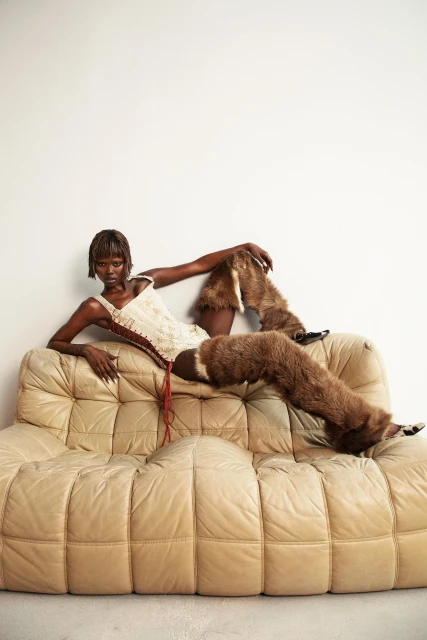 Brunette woman reclines on a tan, tufted sofa wearing a white corset top and furry leg warmers against a white backdrop in a studio.