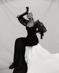 Black and white portrait of a woman in a black gown with sheer polka dot sleeves sitting against a white backdrop.