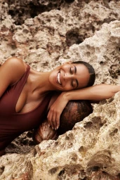Smiling woman in brown swimwear resting her head against textured rocky background with coconuts