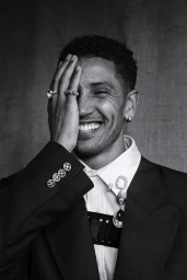 Black and white portrait of a smiling man wearing rings and a blazer covering one eye with his hand.