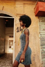 Black woman with afro wearing a denim sleeveless dress standing beside a weathered textured wall