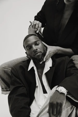 Monochrome portrait of a black man in a black blazer and white shirt with a stylist holding a comb