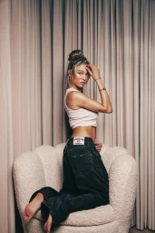 Woman kneeling on a textured armchair wearing a white tank top and baggy dark jeans against pleated drapes