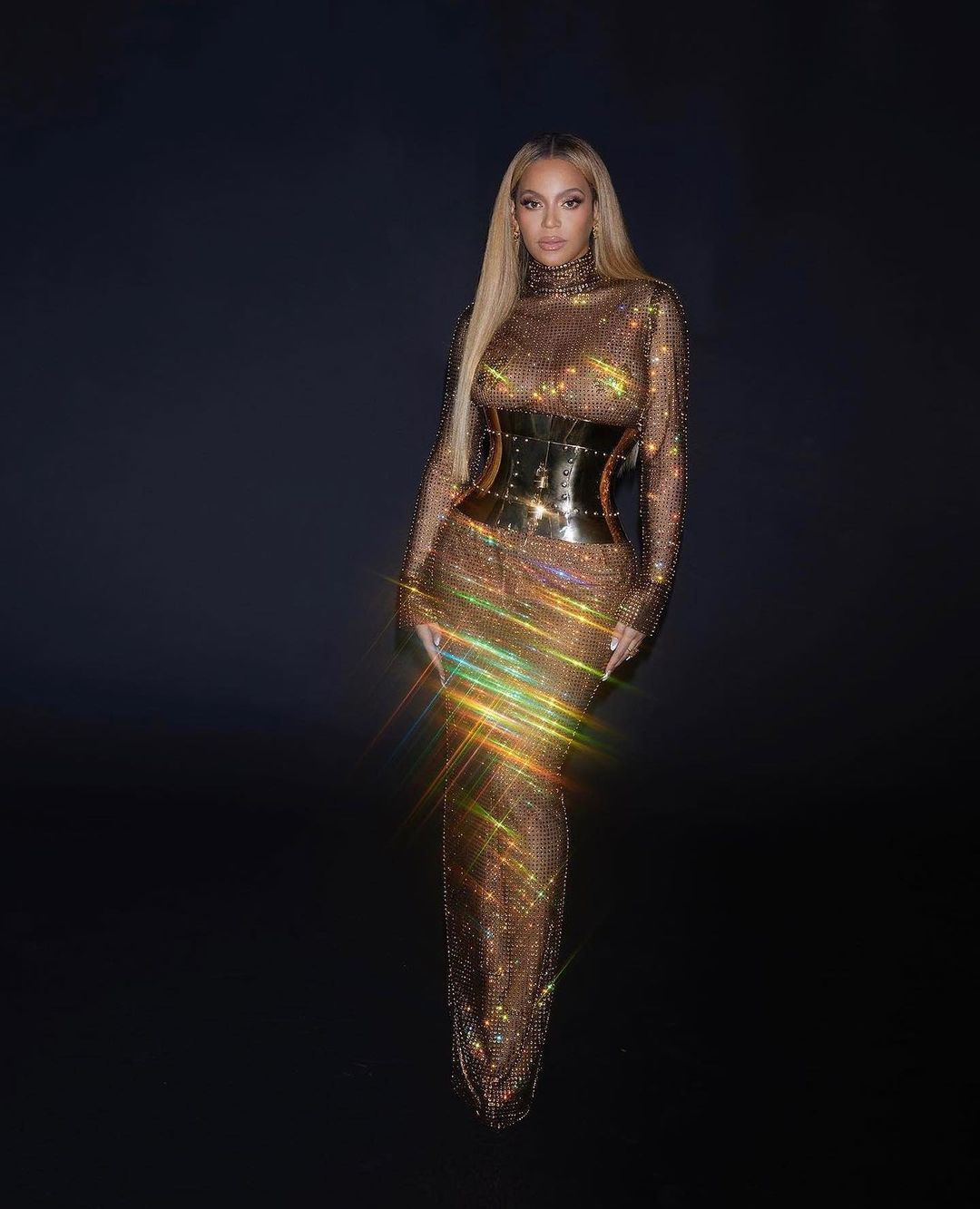 Beyonce poses in a long gold dress with a black waist corset against a dark backdrop, radiating elegance and glamour in a studio setting.