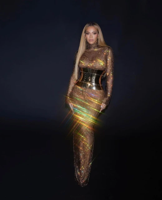 Beyonce poses in a long gold dress with a black waist corset against a dark backdrop, radiating elegance and glamour in a studio setting.