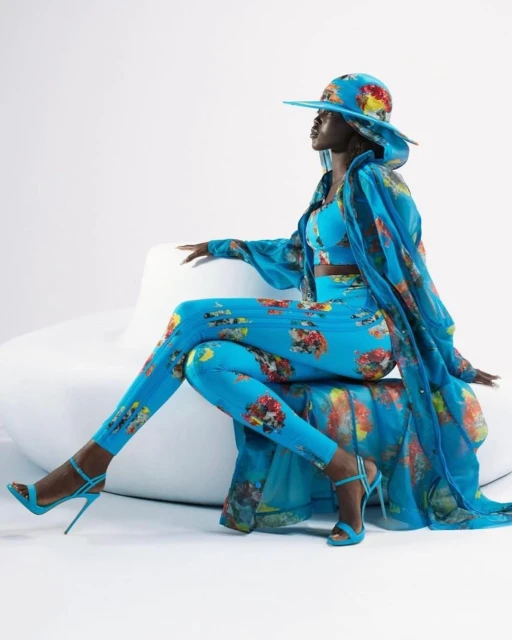 A dark-skinned woman sits elegantly wearing a coordinated aqua floral-print hat, top, leggings, heels, and sheer coat against a white backdrop.