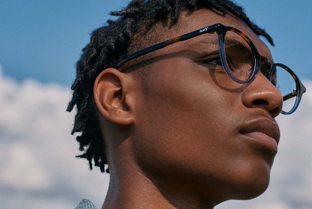 Eye-level close-up of African-American male model with dark curly hair wearing Levi's glasses against a sunny blue sky background.
