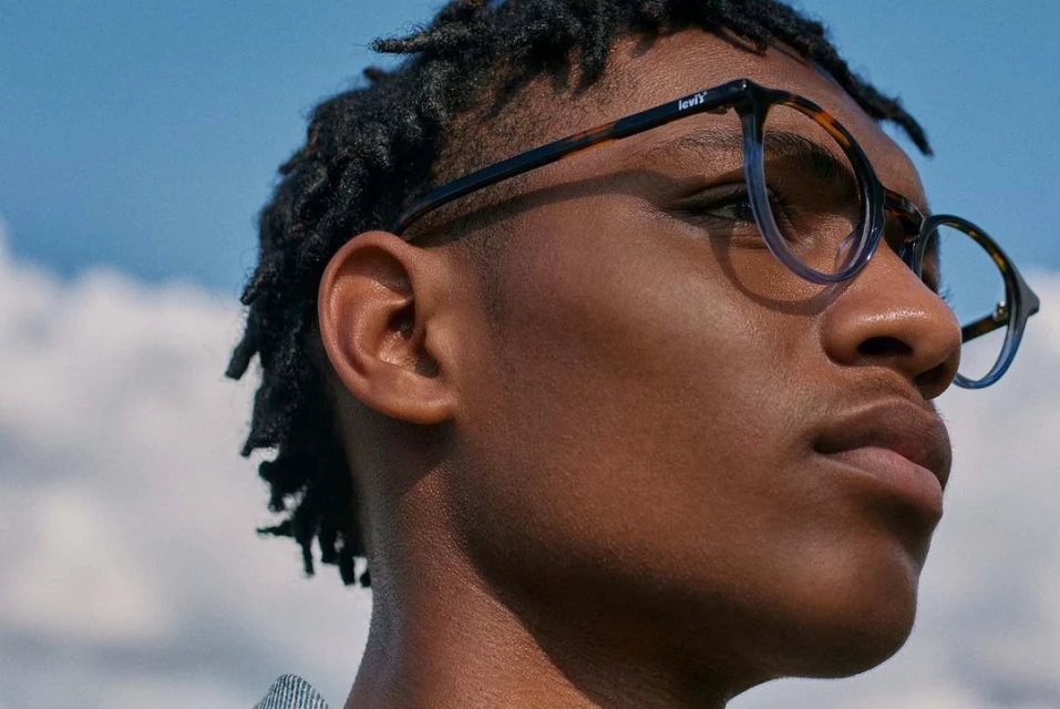 Eye-level close-up of African-American male model with dark curly hair wearing Levi's glasses against a sunny blue sky background.