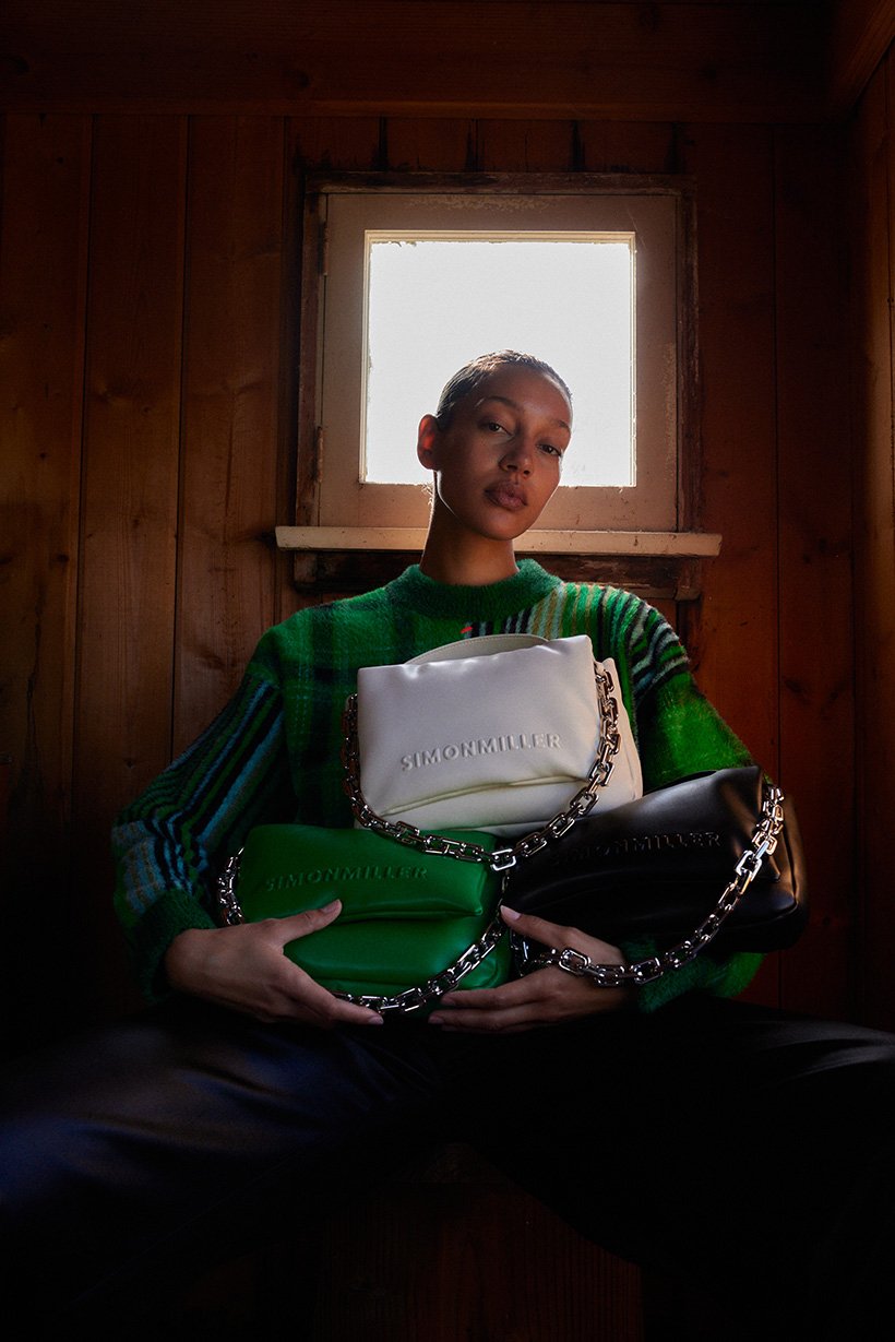 A young adult woman sits in a wooden interior, holding up a green, white, and black purse with silver chain straps and "SIMONMILLER" printed on each.