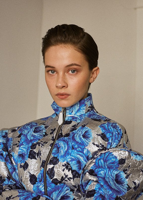 Close-up portrait of a young woman in a blue floral jacket with silver accents, posing in front of a plain, neutral background.