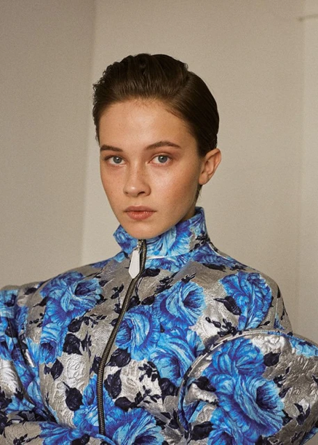 Close-up portrait of a young woman in a blue floral jacket with silver accents, posing in front of a plain, neutral background.
