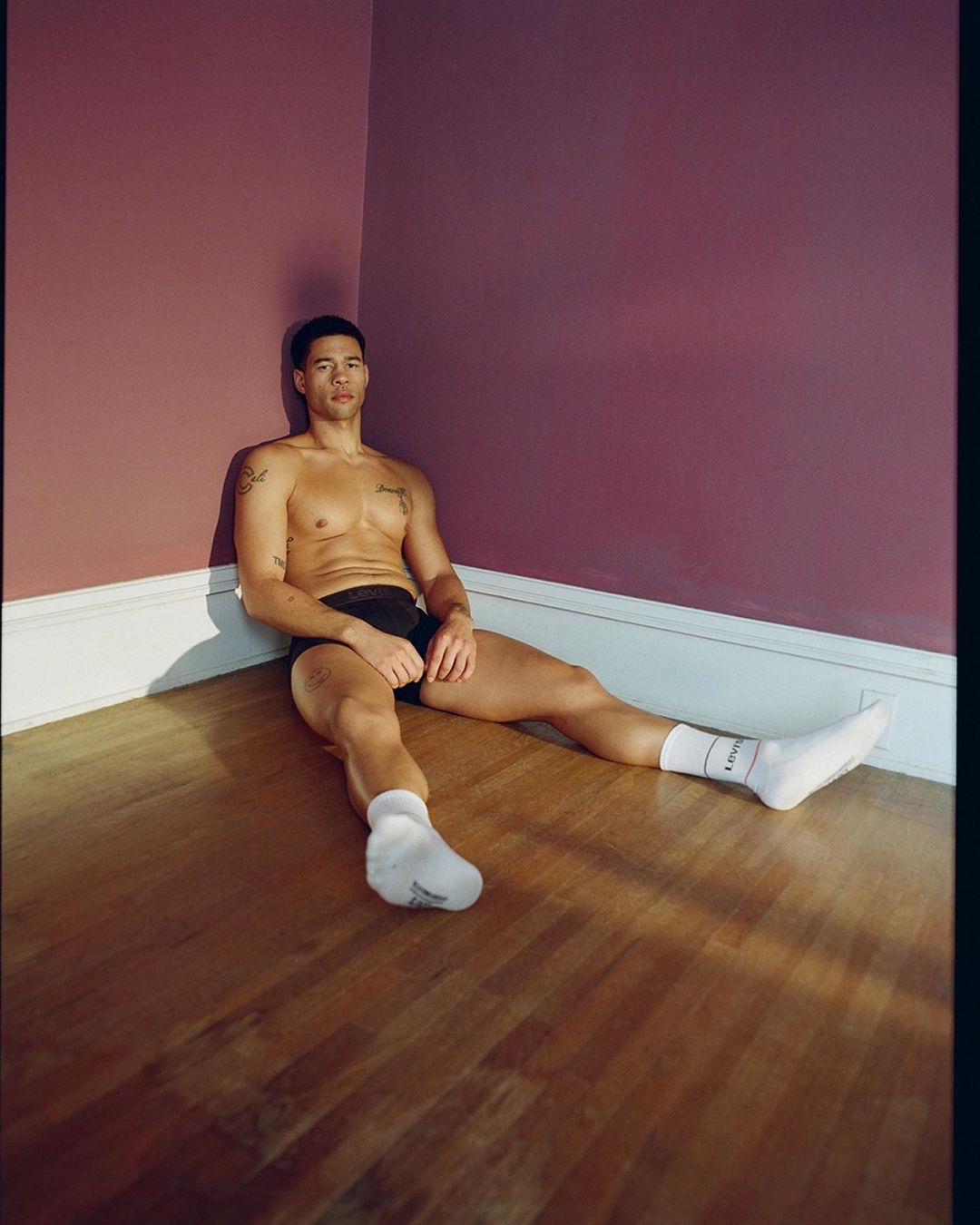 A muscular man in underwear and socks sits in the corner of a room with mauve walls and a wood floor, bathed in natural light.