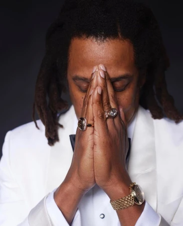 Portrait of a man with dreadlocks in a white tuxedo, eyes closed, hands clasped together near his face, adorned with rings and a gold watch.