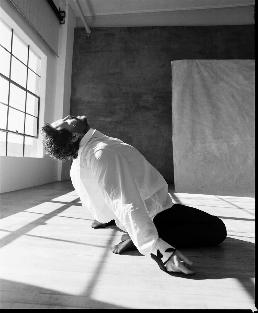 Barefoot male in black pants and a white shirt looks up at the window light, casting shadows on the wood floor and grey walls.