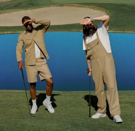 Two men stand on a golf course, shielding their eyes from the sun while holding golf clubs, with a lake and green hills in the background.